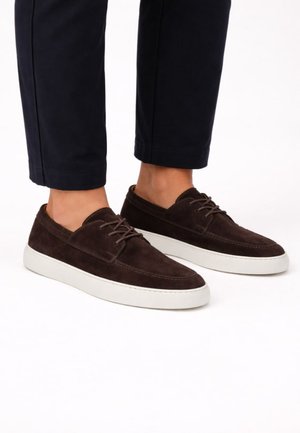 Persoon draagt donkerbruine suède vetersneakers met witte zolen en zwarte cropped broek tegen een witte achtergrond.