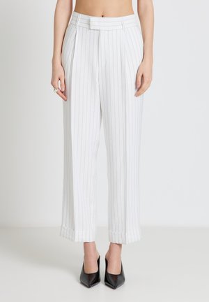 Femme portant un pantalon large blanc à rayures fines, ourlets retroussés, et des escarpins noirs à bout pointu, se tenant devant un fond uni.