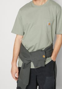 Olijfgroene katoenen t-shirt met een borstzak en Carhartt-logo, gecombineerd met een donkergroen sweatshirt dat om de taille is geknoopt.