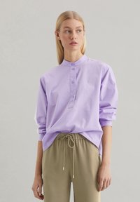 Camicia a maniche lunghe viola chiaro con colletto alzato, patta dei bottoni e vestibilità ampia, abbinata a pantaloni beige con cordoncino.