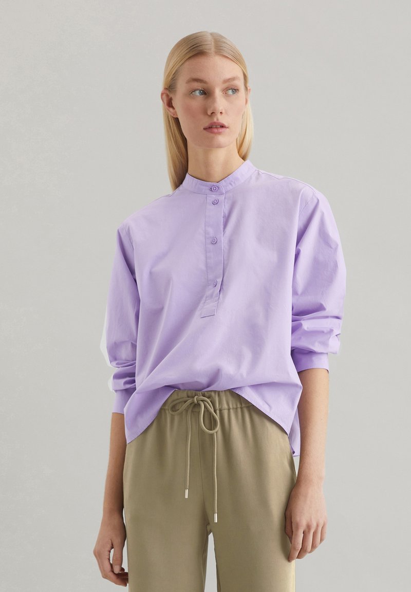 Camicia a maniche lunghe viola chiaro con colletto alzato, patta dei bottoni e vestibilità ampia, abbinata a pantaloni beige con cordoncino.