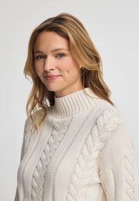 Cable-stickad polotröja i krämvit. Har tjock ribbad krage, texturerade mönster och intrikata flätor. Mjuk stickad tyg.