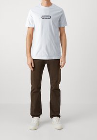 Muž v bílém tričku s grafikou s nápisem "Hilfiger New York City 1985", tmavě hnědých kalhotách a bílých teniskách, stojící na jednobarevném pozadí.