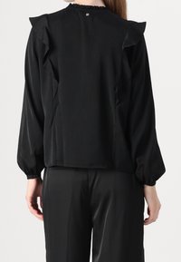 Haut noir à manches longues avec des accents à volants sur les épaules, un col froncé et une petite fermeture à bouton à l'arrière. Tissu à texture lisse.