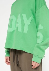 Sudadera corta verde con letras en negrita "DAY", tejido texturizado, dobladillo sin rematar y puños acanalados. Combinada con pantalones marrones.
