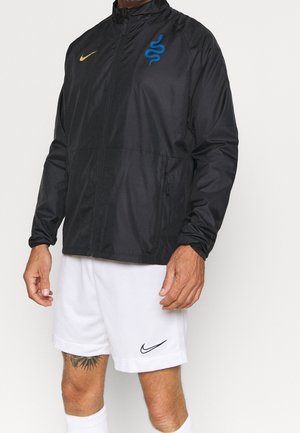 Uomo con giacca nera con cerniera e logo serpente blu e pantaloncini bianchi Nike con swoosh nero, in piedi davanti a uno sfondo semplice.