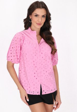 Helles rosa Kurzarmbluse mit Lochmuster, Knopfleiste und Stehkragen, kombiniert mit schwarzen Shorts.