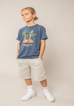 Garçon les mains dans les poches portant un t-shirt bleu "Tropical Club", un short beige, des chaussettes blanches et des baskets blanches, debout devant un fond uni.