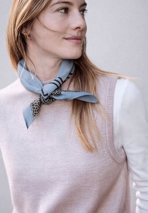 Žena se světle hnědými vlasy, která má na sobě světlý růžový svetřík bez rukávů přes bílý dlouhý rukáv a modrý vzorovaný šátek na krk.