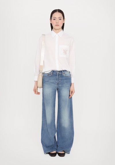Camisa blanca de manga larga con un logo en el bolsillo, combinada con unos vaqueros de pierna ancha azul claro y zapatos oscuros de estilo slip-on, de pie frente a un fondo liso.