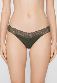 Dunkelgrüner String mit einem zarten Spitzenbund. Verfügt über eine gerippte Textur am Körper und florale Spitzenverzierungen am oberen Rand.