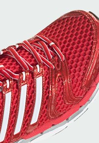 Chaussure de course en mesh rouge avec des lacets rayés blancs et rouges et des bandes blanches sur le côté au-dessus d’une semelle rouge et blanche texturée.