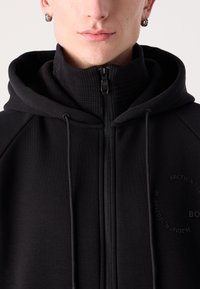 Sweat à capuche zippé noir avec col montant, tissu texturé, cordons de serrage et détail de logo embossé sur le côté gauche. Design lisse et ajusté.