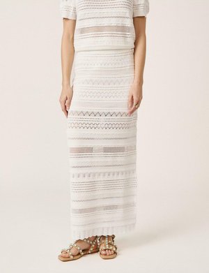 Falda de punto de crochet blanca con patrones geométricos, combinada con un top de manga corta a juego y sandalias planas beige con tachuelas.