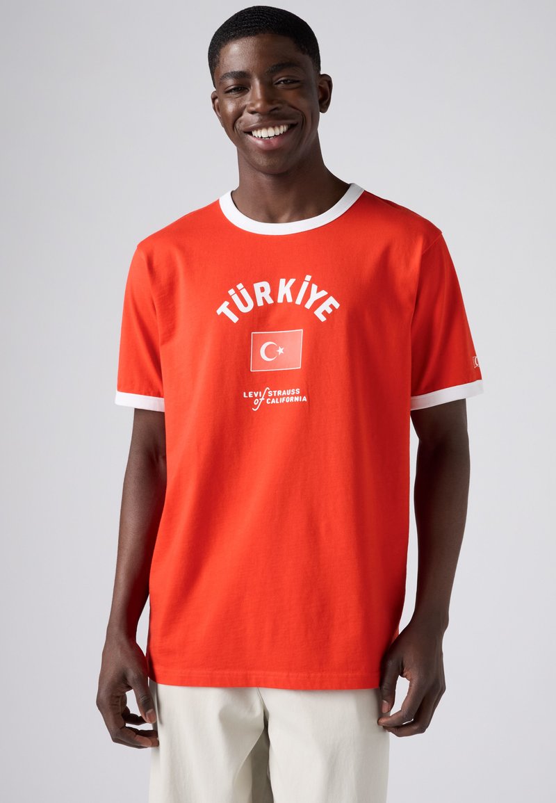 Young man smiling, wearing a red T-shirt with white trim featuring "TÜRKİYE," a Turkish flag, and Levi's branding on front, paired with light pants.