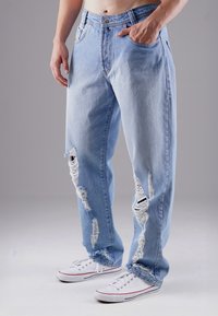 Hellblaue, ausgewaschene Jeans mit großen Rissen, die ausgefranste Kanten zeigen. Sie verfügen über ein klassisches Fünf-Taschen-Design und eine abgenutzte Textur, kombiniert mit weißen Sneakers.