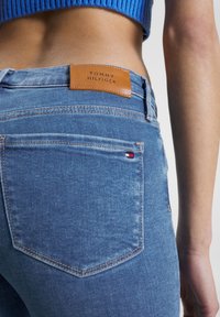 Svetle modré džíny z denimu s koženou visačkou Tommy Hilfiger na pase a malým vlajkovým logem na zadní kapse. Pohled zblízka.