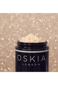 Bocal noir étiqueté "OSKIA LONDON" rempli de poudre légère et granuleuse, avec un bord lisse et une finition brillante. L'arrière-plan présente un flou bokeh doux.