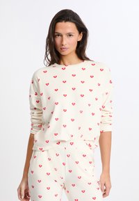 Vaatteet vapaamuotoiseen oleskeluun, vaalea kreemi väri, joissa on hajallaan punaisia sydänkuvioita; löysä malli, pitkät hihat ja joustava vyötärö housuissa.