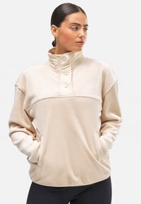 Sudadera de forro polar beige con un cuello alto, tapeta de botones y bolsillos laterales. Textura suave; corte holgado; diseño limpio; mangas largas.