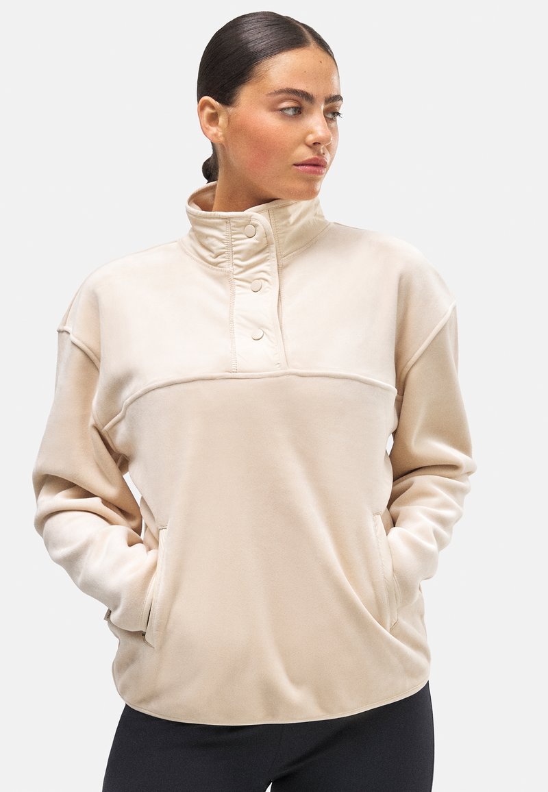 Sudadera de forro polar beige con un cuello alto, tapeta de botones y bolsillos laterales. Textura suave; corte holgado; diseño limpio; mangas largas.
