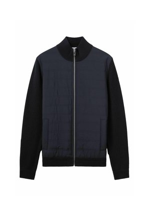 Veste bleu marine foncé avec un col et des poignets côtelés. Présente un panneau avant matelassé et une fermeture éclair. Deux poches latérales. Manches en maille texturée.