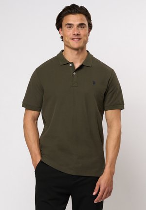 Bărbat purtând tricou polo cu mânecă scurtă, verde măsliniu, stând cu o mână în buzunar, pe un fundal simplu, deschis la culoare.