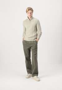 Maglione di lana a trecce di colore crema con colletto a zip, abbinato a pantaloni larghi color verde oliva e sneaker off-white. Design semplice e texturizzato.