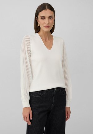 Pull blanc à col en V avec des manches longues texturées et côtelées. Porté avec un jean en denim foncé ; textile lisse, coupe décontractée.