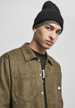 Jaunas šviesios odos vyras dėvi juodą megztą kepurę ir alyvuogių žalios spalvos zomšinę striukę ant juodos palaidinės, žiūri šiek tiek į šoną.