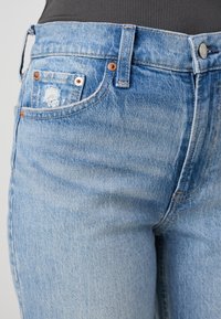 Světle modré džínové kalhoty s vybledlým povrchem, vybavené měděnými detaily, přední kapsou a odřenými prvky v okolí kapsy.