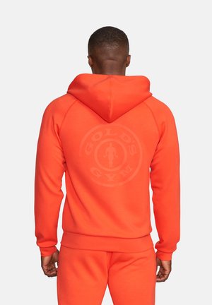 Mann von hinten gesehen, trägt leuchtend orangefarbenen Kapuzenpullover und Jogginghose mit schwachem Gold's Gym-Logo auf der Rückseite des Kapuzenpullovers.