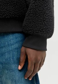 Schwarzer Fleece-Pullover mit einer strukturierten, flauschigen Oberfläche; im Hintergrund sind ein gerippter schwarzer Bund und blaue Jeans zu sehen.