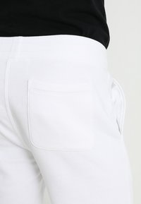 Białe spodnie dresowe z prążkowanym pasem i jedną tylną kieszenią. Materiał wydaje się miękki i teksturowany, odpowiedni do noszenia na co dzień.