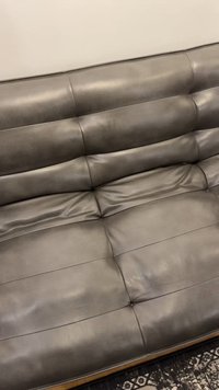 Šedá kožená pohovka s prošívaným opěradlem a sedáky, umístěná na vzorovaném koberci u světle zbarvené zdi.