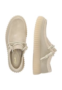 Zapatos de ante y lona beige con una suela de goma texturizada, que presentan costuras decorativas y un diseño con cordones. Marca Clarks visible en el interior.
