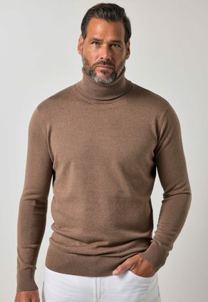 Man met baard die een bruine coltrui en witte broek draagt, staat met één hand in zijn zak tegen een effen achtergrond.