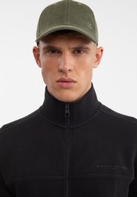 Giacca nera con zip in tessuto testurizzato, abbinata a un cappello in velluto a coste verde oliva con cuciture a contrasto e un piccolo logo.