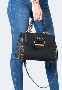 Personne en jean bleu tenant un sac à main en cuir noir avec des clous et des ornements dorés par ses poignées supérieures.