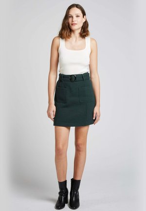 Jupe mini en laine verte avec deux poches avant, taille ceinturée avec une boucle ronde, associée à un débardeur en maille blanc et des bottines noires.