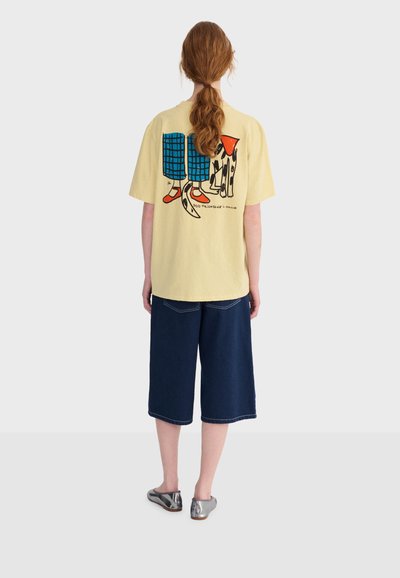 Femme aux cheveux roux attachés en queue de cheval, portant un t-shirt jaune avec un motif de chien et de pattes, un short large bleu marine, et des ballerines argentées métalliques, vue de dos.