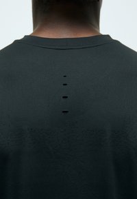 T-shirt de sport noir en tissu texturé, avec un col rond et cinq découpes horizontales en forme de bouton le long du dos.