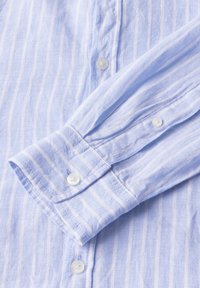 Lichtblauwe linnen blouse met witte verticale strepen, knoopmanchetten en kleine, doorzichtige knopen. De textuur lijkt zacht en iets gekreukt.