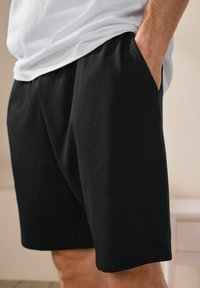 Black cotton shorts with an elastic waistband, side pockets, and a relaxed fit. The fabric has a smooth, soft texture.