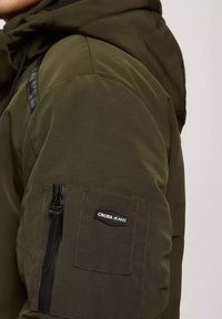 Oliwkowa kurtka z kapturem z zamkiem błyskawicznym na rękawie, ozdobiona czarną metką "CROSS JEANS" i teksturowanym materiałem.