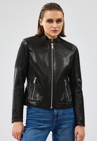 Chaqueta de cuero negra con un alto cuello, acentos acanalados en los hombros, cierre delantero y dos bolsillos laterales. Llevada sobre vaqueros azules.