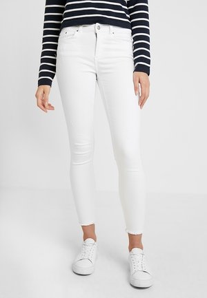 Frau trägt weiße Skinny-Jeans mit ausgefransten Säumen, weiße Turnschuhe und ein schwarz-weiß gestreiftes Langarmshirt, steht vor einem einfarbigen Hintergrund.