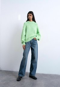 Femme portant un pull oversize vert clair, un jean bleu à jambes larges et des chaussures noires, debout devant un mur blanc uni.