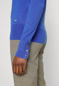 Blauer Strickpullover mit strukturierten Handgelenkfesseln und drei dekorativen Knöpfen. Kombiniert mit gemusterten karierten Hosen.