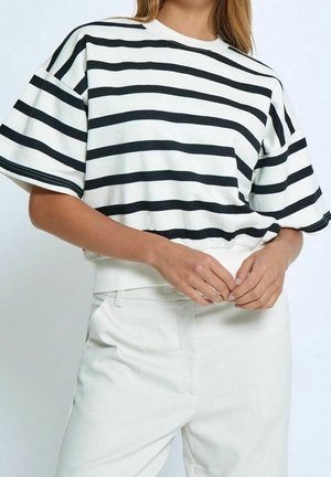 Kobieta ubrana w biały krótki t-shirt w czarne poziome paski oraz białe spodnie z wysokim stanem, z luźno splecionymi rękami na wysokości talii.